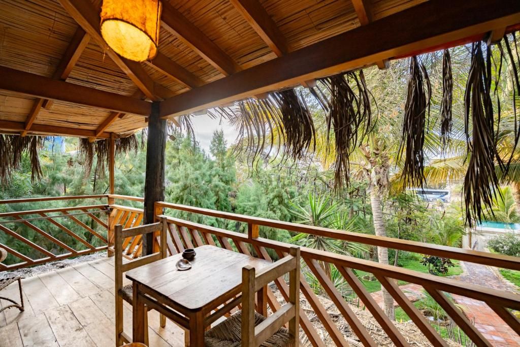 Una terraza de madera con una mesa y sillas, rodeada por una barandilla, que da vista a un jardín exuberante con varios árboles y una piscina.