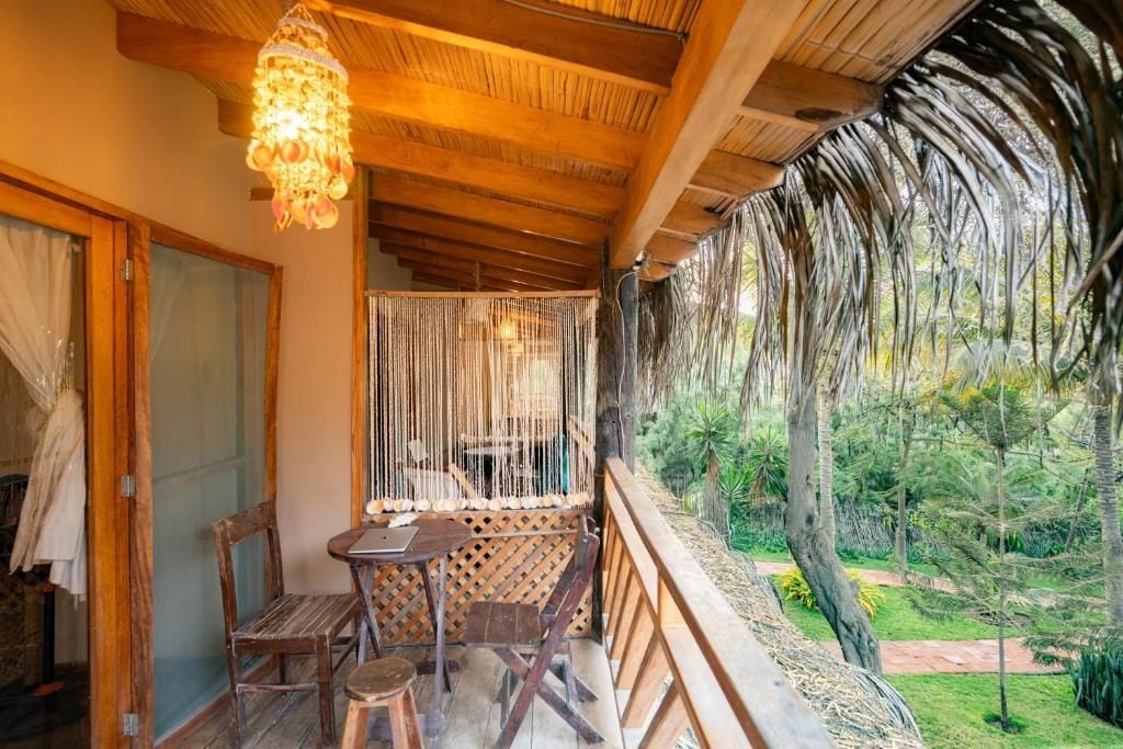 Un balcón de madera con una mesa y sillas, un colgante de luz y vistas a un jardín con árboles.