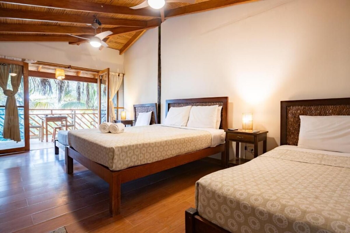 A hotel room with two beds, wooden flooring, a ceiling fan, and a balcony with a view.