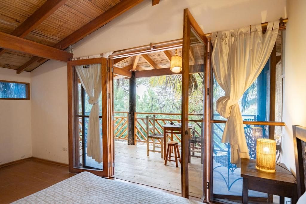 Una habitación con techo de madera, una cama, un ventanal con cortinas y vistas a una terraza exterior con mesa y sillas.