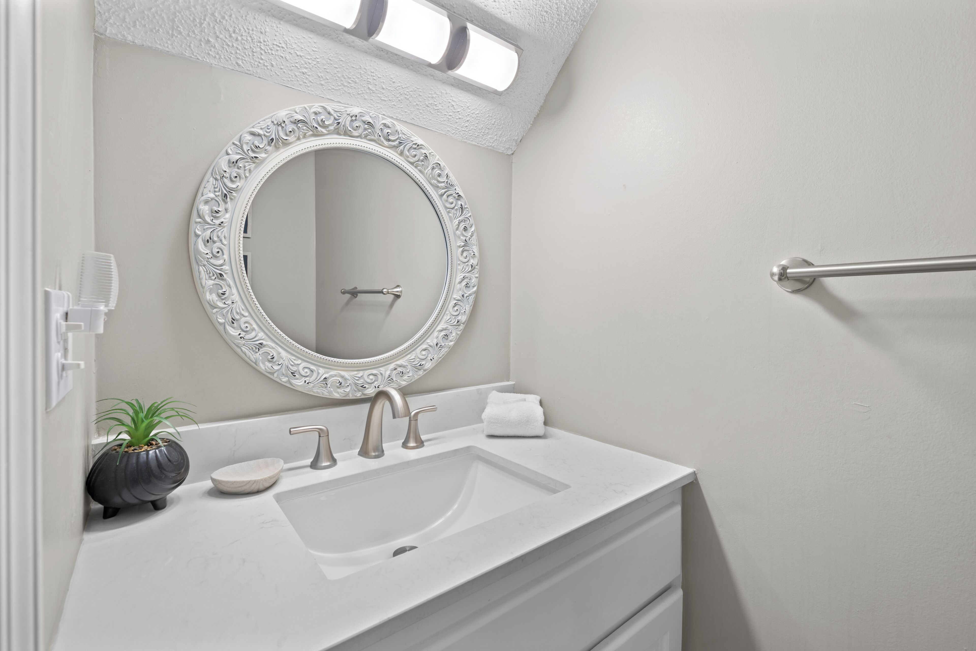 A bathroom with a white vanity, a round mirror with ornate detailing, a towel, and a potted plant.