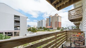 Una terraza de madera con mobiliario exterior, con vista a un estacionamiento y un edificio de varios pisos.