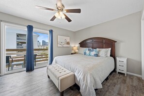 A bedroom with a bed, a nightstand, a bench, a ceiling fan, and a view of a balcony with a railing. - Gulf View 8 (Gulf Shores)