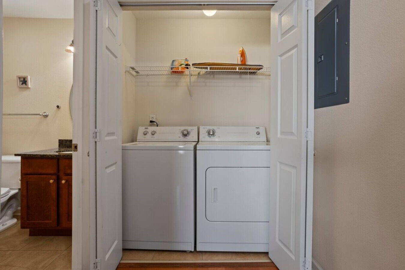 A laundry room with two white washers and dryers, a shelf with a bottle and a box, and a wooden cabinet.