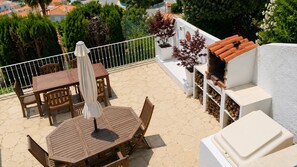 Un patio con una mesa y sillas de madera, un paraguas blanco y una pared blanca con sección de techo de tejas.