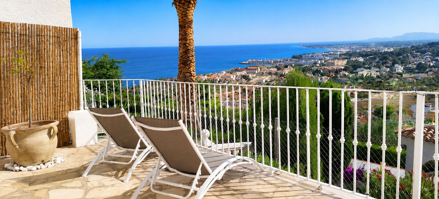 A balcony with two sun loungers, a potted plant, and a view of a coastal town.