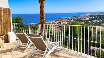 A balcony with two sun loungers, a potted plant, and a view of a coastal town.