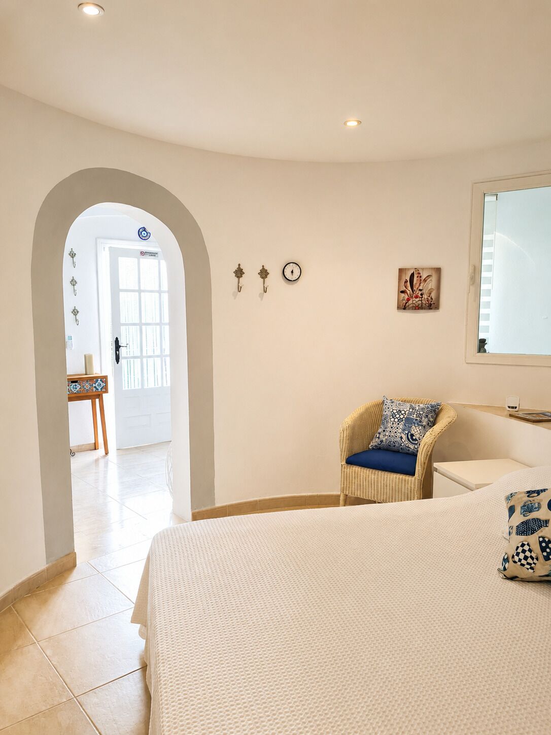 Une chambre d’hôtel avec un lit, une chaise avec un coussin, une petite table, une horloge murale et un tableau.