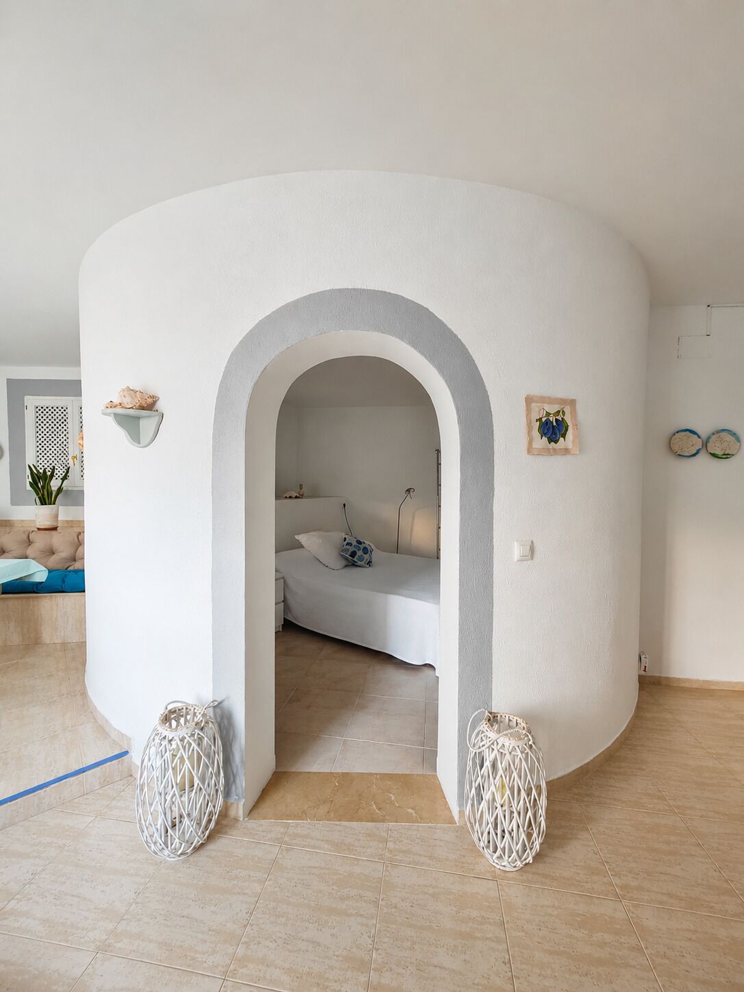 Een kamer met een gebogen witte muur, een bed met een witte matras en een blauw kussen, een klein wit plankje met decoratie, en twee witte gevlochten manden.