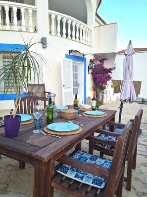 Een terras met een houten tafel voor twee personen, omringd door rieten stoelen met blauwe kussens.