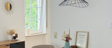 A dining area with a wooden table, chairs, a hanging light fixture, a window with a view of greenery, and a wall-mounted mirror.