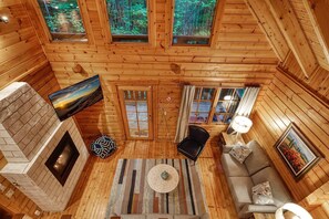 A log cabin interior with a fireplace, a sofa, a dining table, and a view of the forest through the windows. - Romantic Cabin | Sauna & Hot Tub near Dollywood (Sevierville)