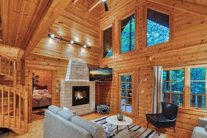 A log cabin interior with a fireplace, a sofa, a dining table, and a large window offering a view of the forest. - Romantic Cabin | Sauna & Hot Tub near Dollywood (Sevierville)