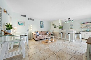 A modern living room with a glass dining table, a sofa, and a kitchen area with white cabinets and a hanging light fixture. - SummerSalt - 5 Min Walk To Beach! (Jupiter)