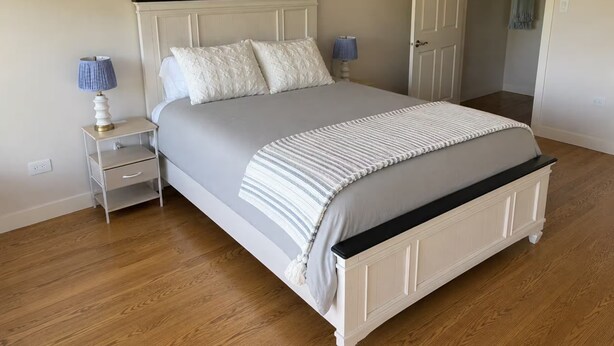 Un dormitorio con cama, mesitas de noche, una lámpara y una puerta que da a otra habitación.