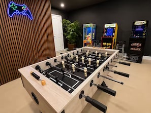 Ein Tischkicker in einem Raum mit leuchtendem Neon-Controller-Schild, Spielautomaten und einer Topfpflanze.