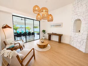 A modern living room with a round wooden coffee table, a sofa, and a dining area with chairs. There is a stone wall with an archway and a large window with a view of outdoor furniture. - Heated Pool, Arcade, Home Theater, Outdoor Dining (Cape Coral)