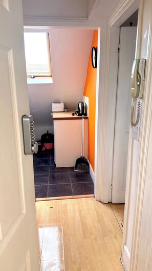 Ein enger Flur, der zu einem Zimmer mit Fenster, einer Tür mit Telefon und einer kleinen Küchenzeile mit Spüle und Wasserkocher führt.
