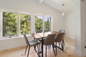 A dining area with a wooden table and chairs, large windows with a view of trees, and a hanging light fixture.