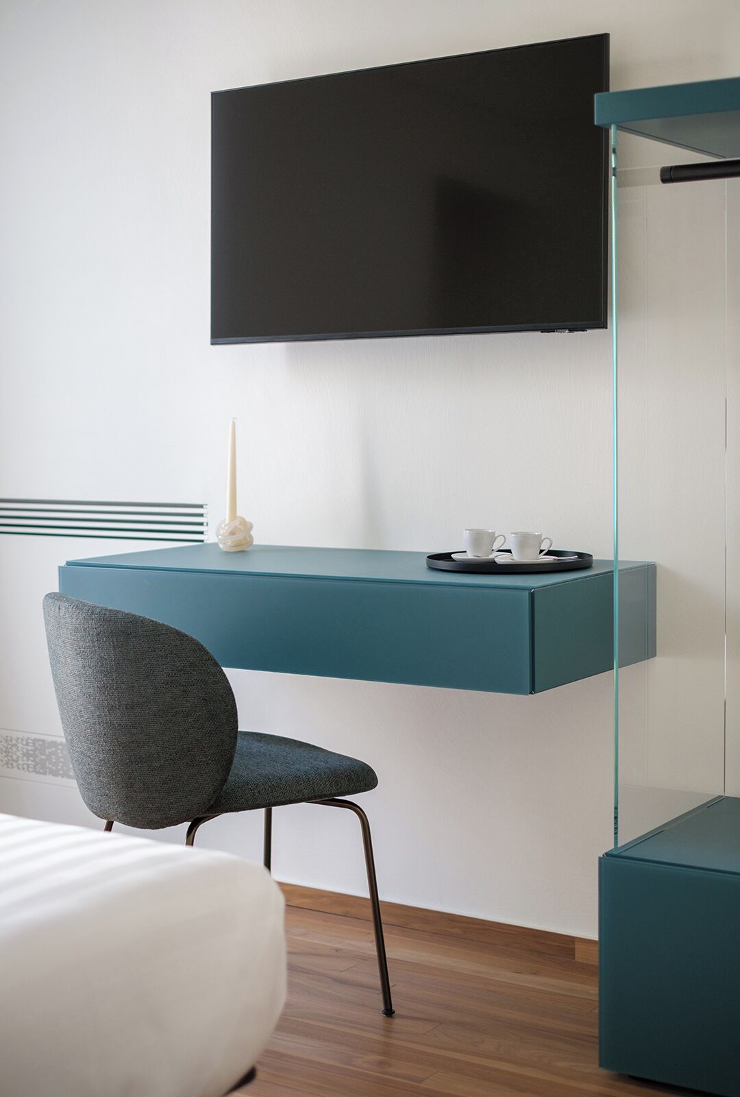 Una camera moderna con una scrivania verde acqua, una sedia, una TV a schermo piatto montata a parete e un comodino con una lampada bianca.