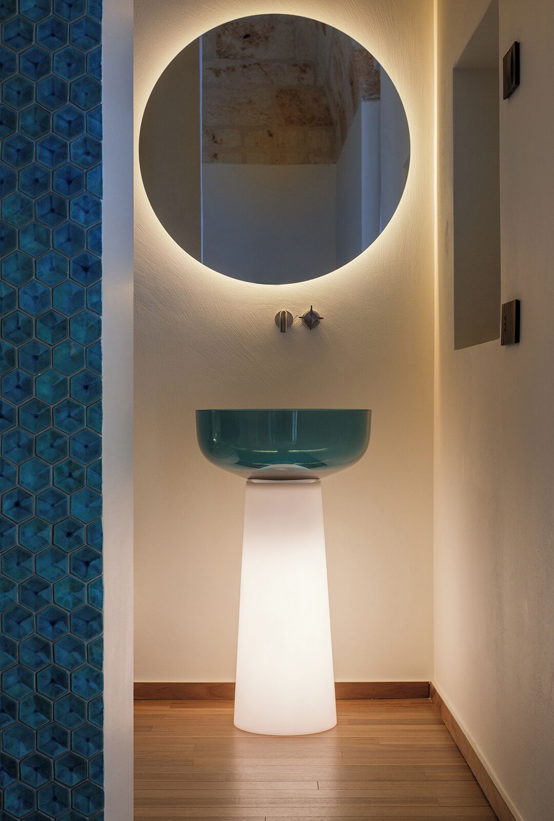 A bathroom with a round mirror, a pedestal sink, and a hexagonal tile pattern on the wall.