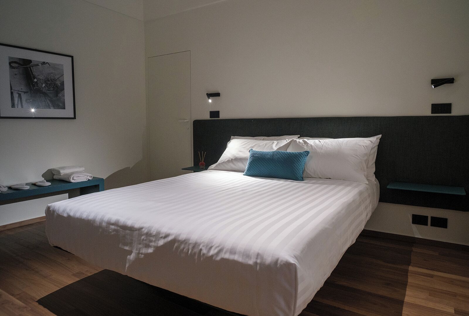 Una cama bien hecha con sábanas blancas y una única almohada azul, una mesita de noche con lámpara, y un cuadro en la pared.