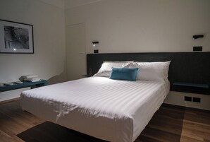 Una cama bien hecha con sábanas blancas y una única almohada azul, una mesita de noche con lámpara, y un cuadro en la pared.