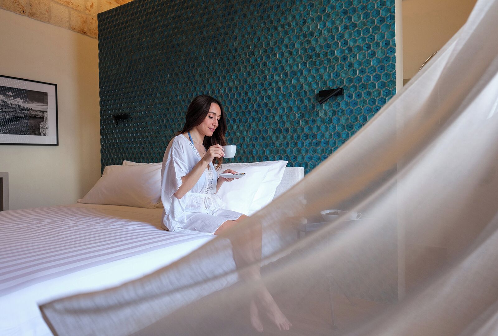 A bed with a white bedspread, a person sitting on the bed holding a cup and a plate, and a blue textured wall behind.