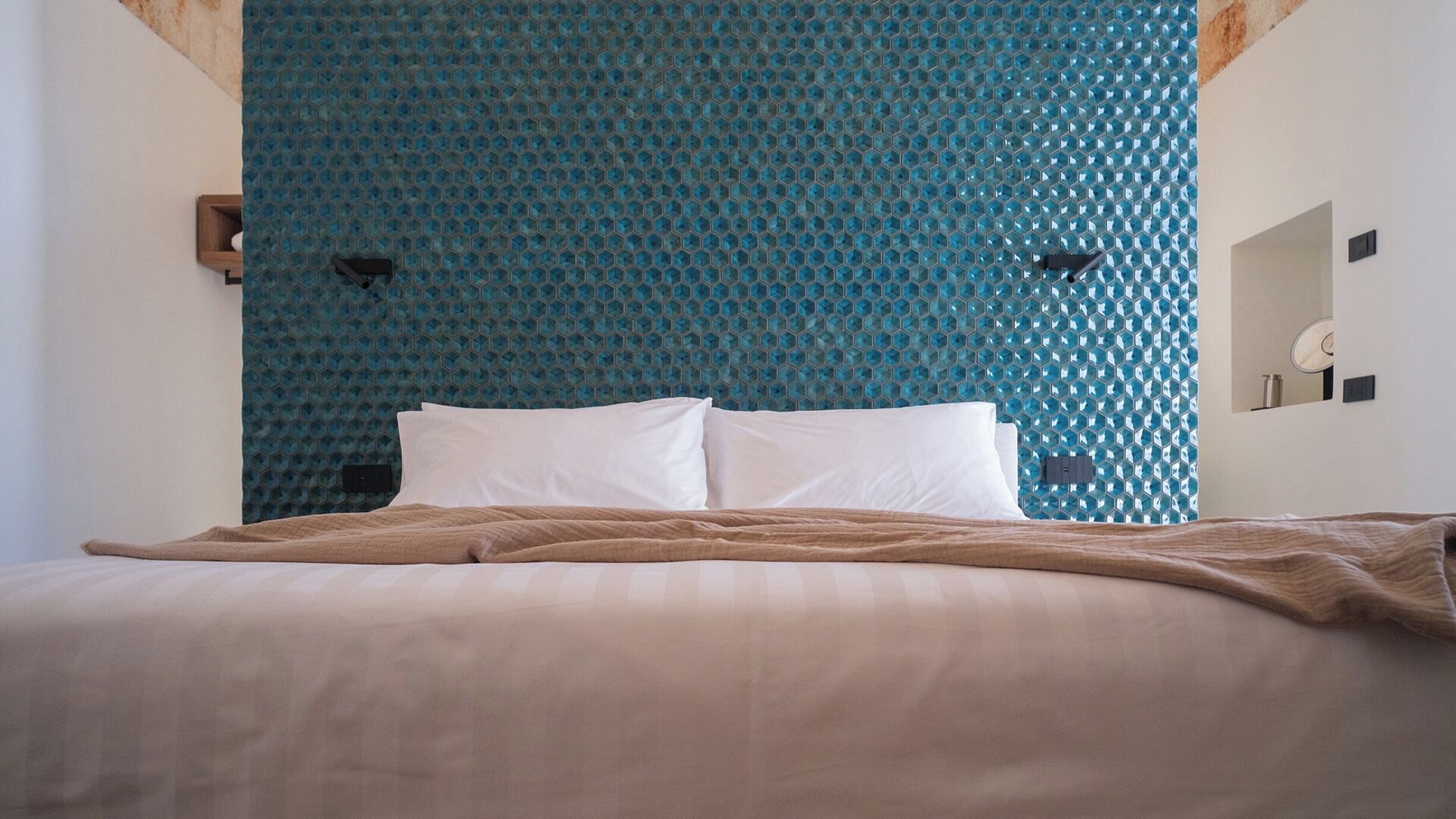 A bed with white pillows and a brown blanket, a blue patterned wall, and a small wooden shelf with a mirror and a light switch.