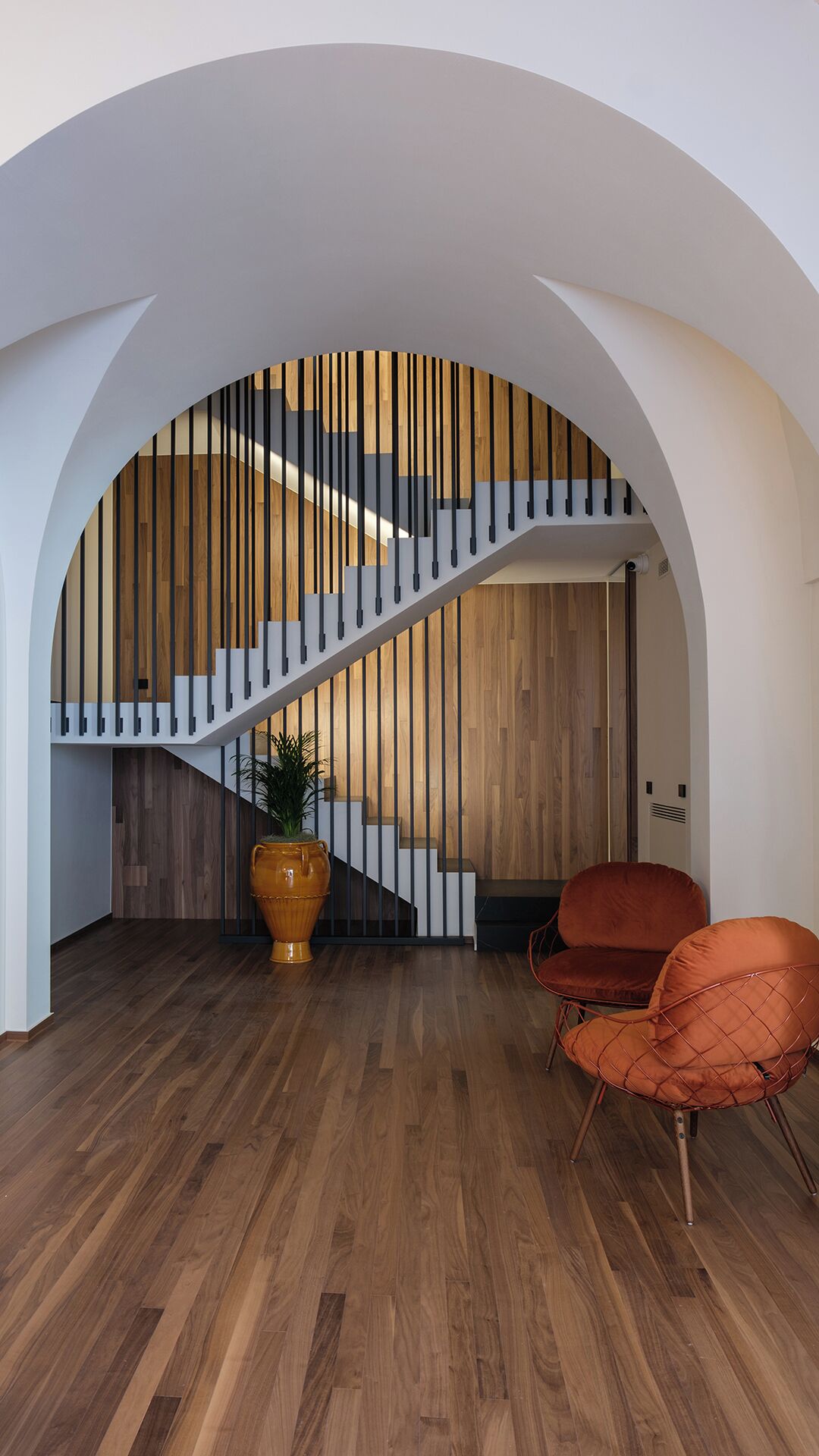 Um interior moderno com uma escada de madeira, um vaso grande e duas cadeiras laranja.