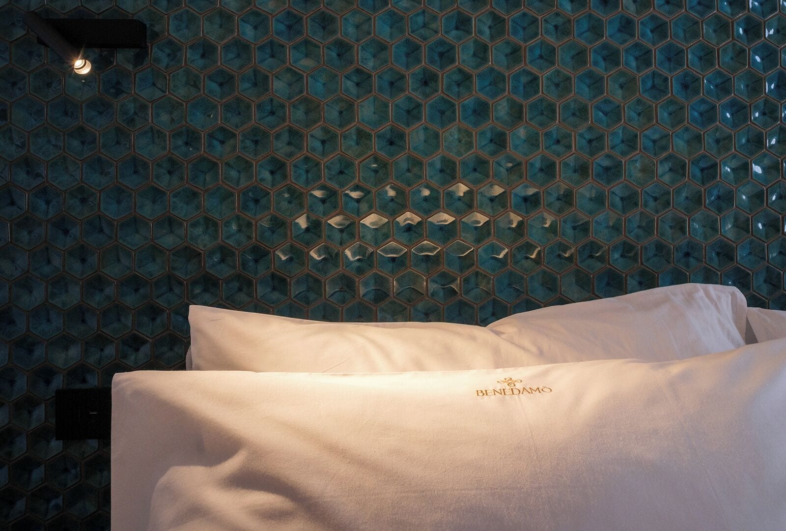 Une chambre d’hôtel avec un mur orné d’un motif hexagonal, un lit avec une literie blanche et un luminaire placé au-dessus du lit.