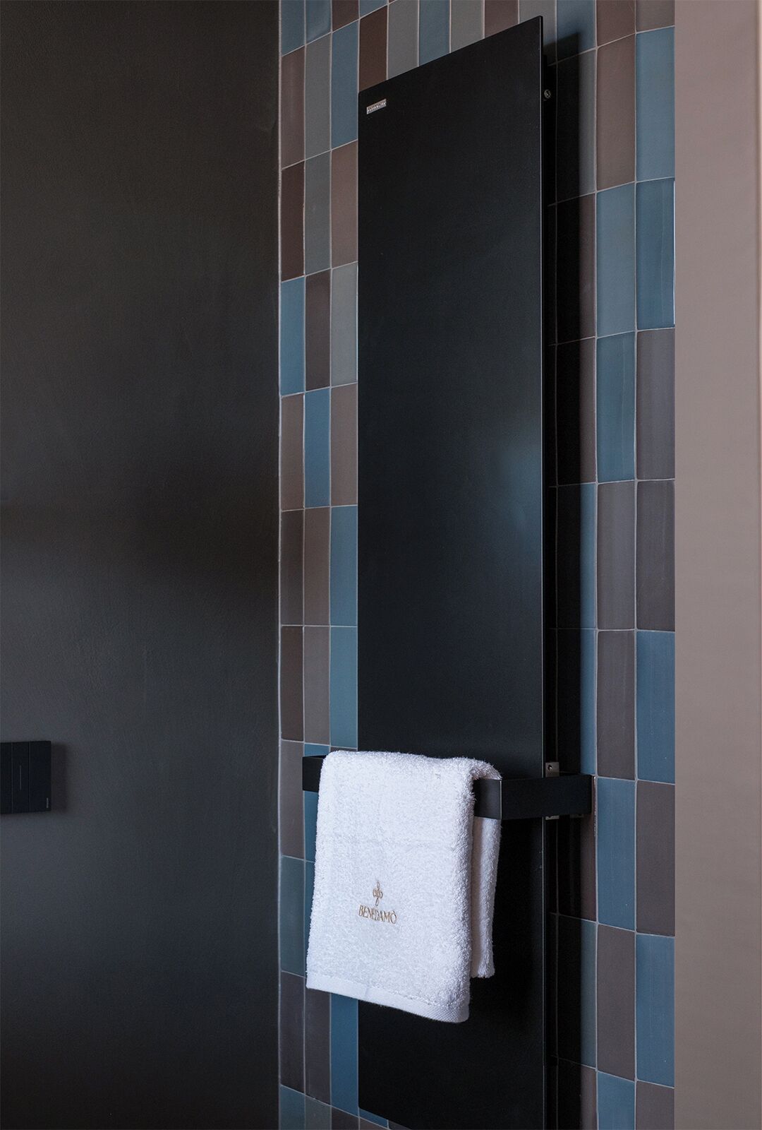 A black towel rack with a white towel hanging on it, set against a tiled wall with a mix of blue, brown, and gray tiles.