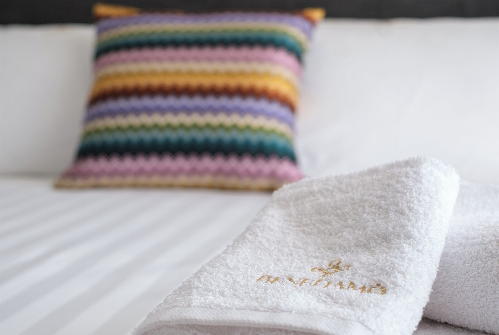 A white bed with a striped pillow and two white towels with embroidered text.