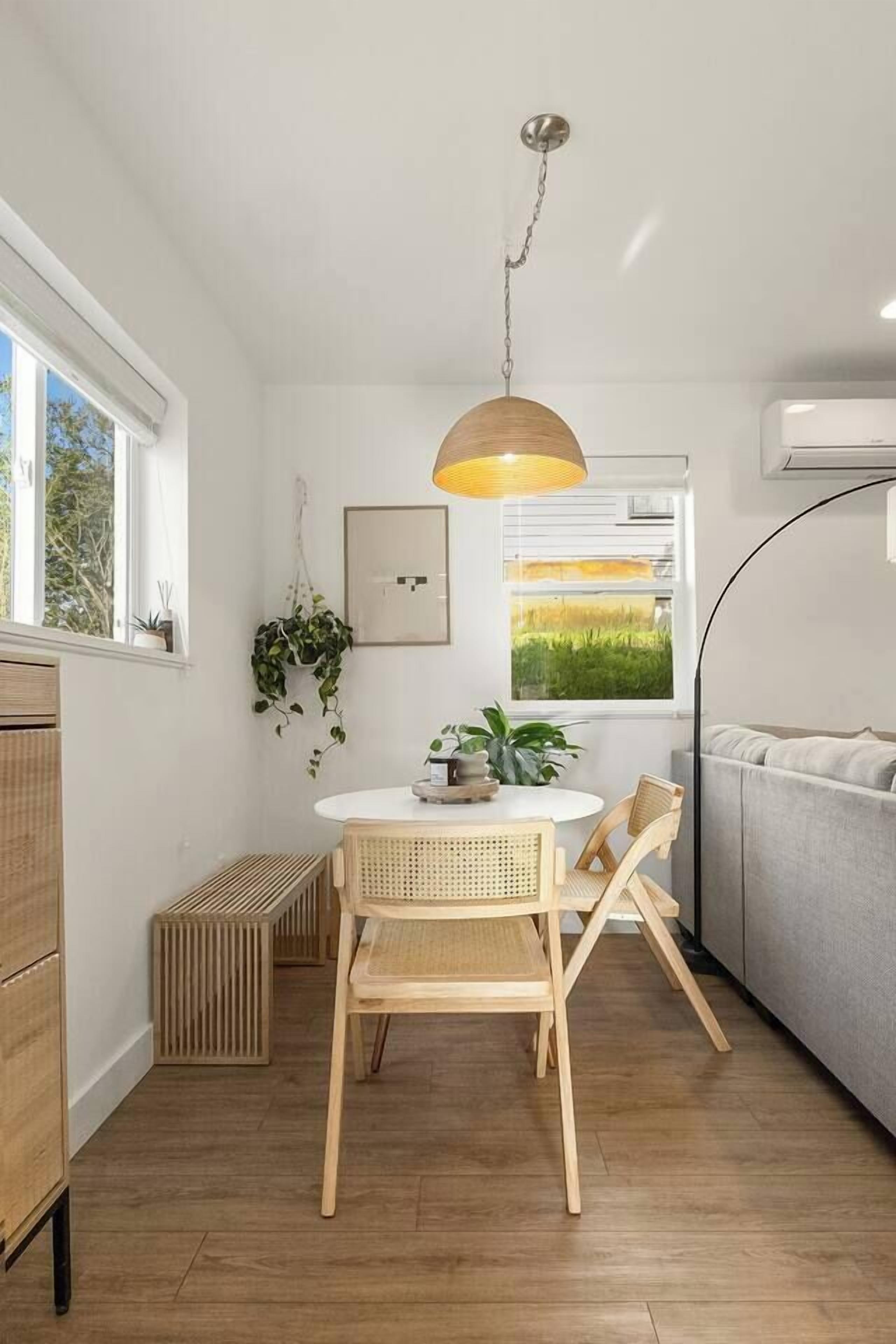 A modern kitchen with a round table, wooden chairs, a hanging light fixture, and a view of greenery through a window.