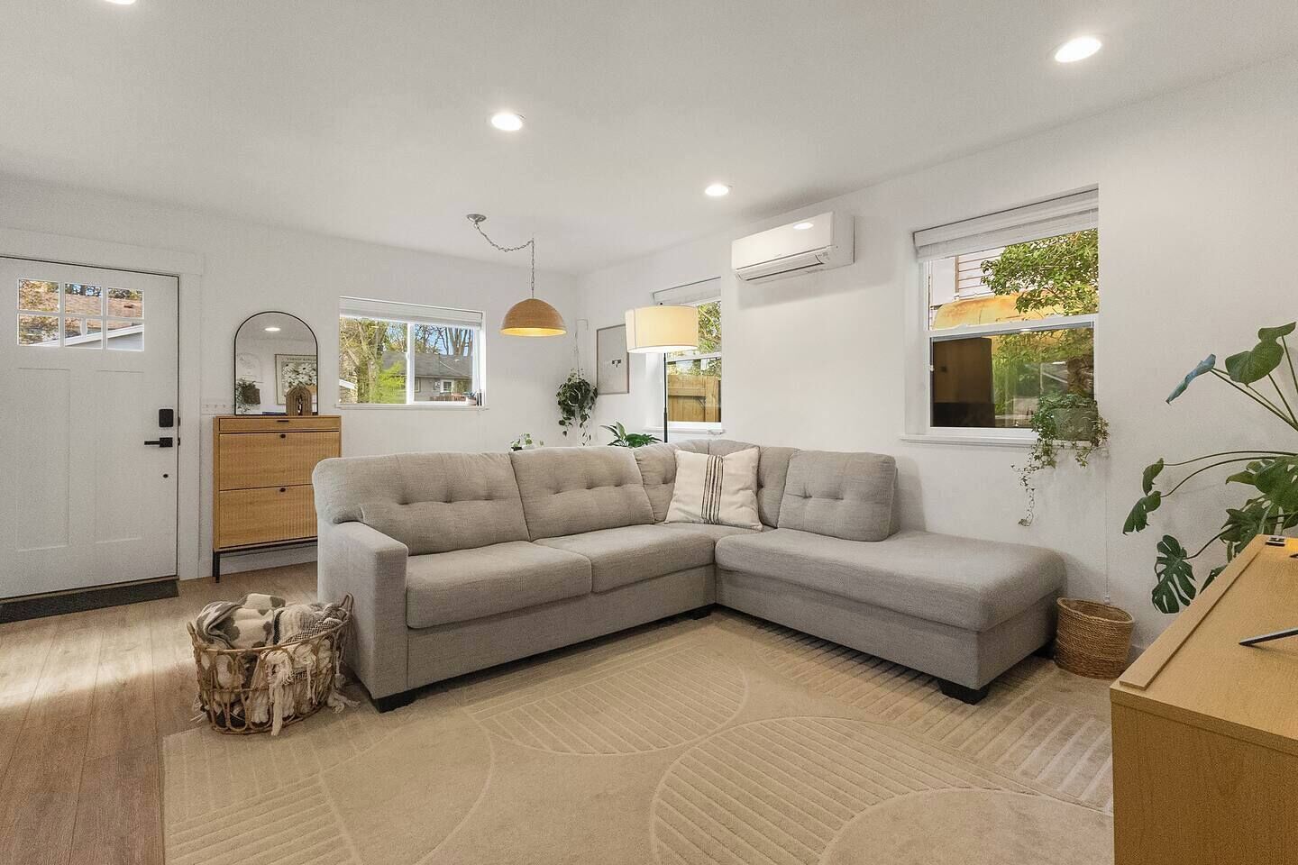 A modern living room with a grey sectional sofa, wooden cabinet, and a large window with a view of greenery.
