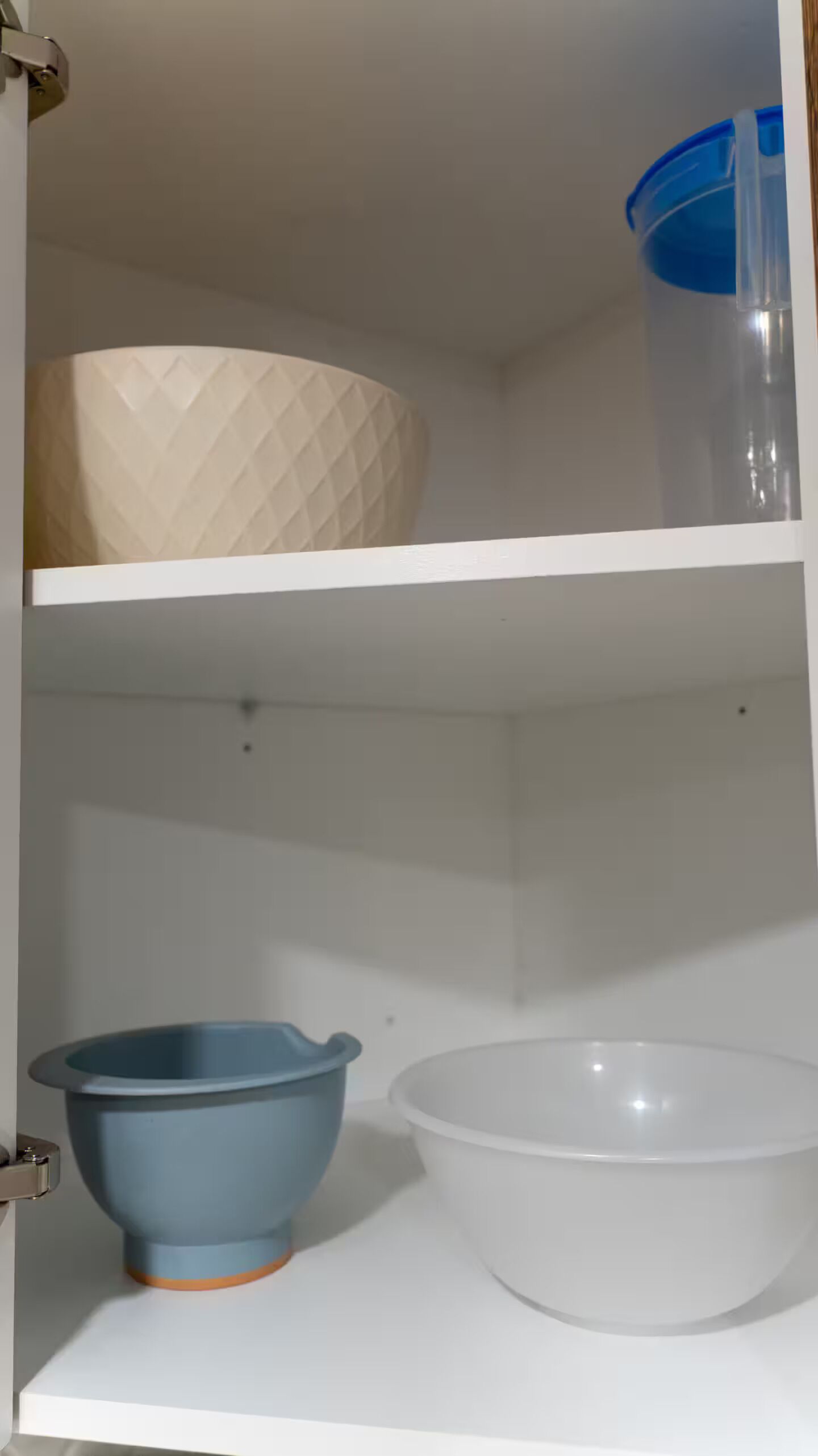 Een keukenkastje met een beige kom met textuur op het bovenste plank, een blauwe kom met handgreep op het middelste plank en een witte kom op het onderste plank.