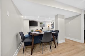Un espace repas moderne avec une table et des chaises en bois, un téléviseur à écran plat fixé au mur et une fenêtre avec des stores.