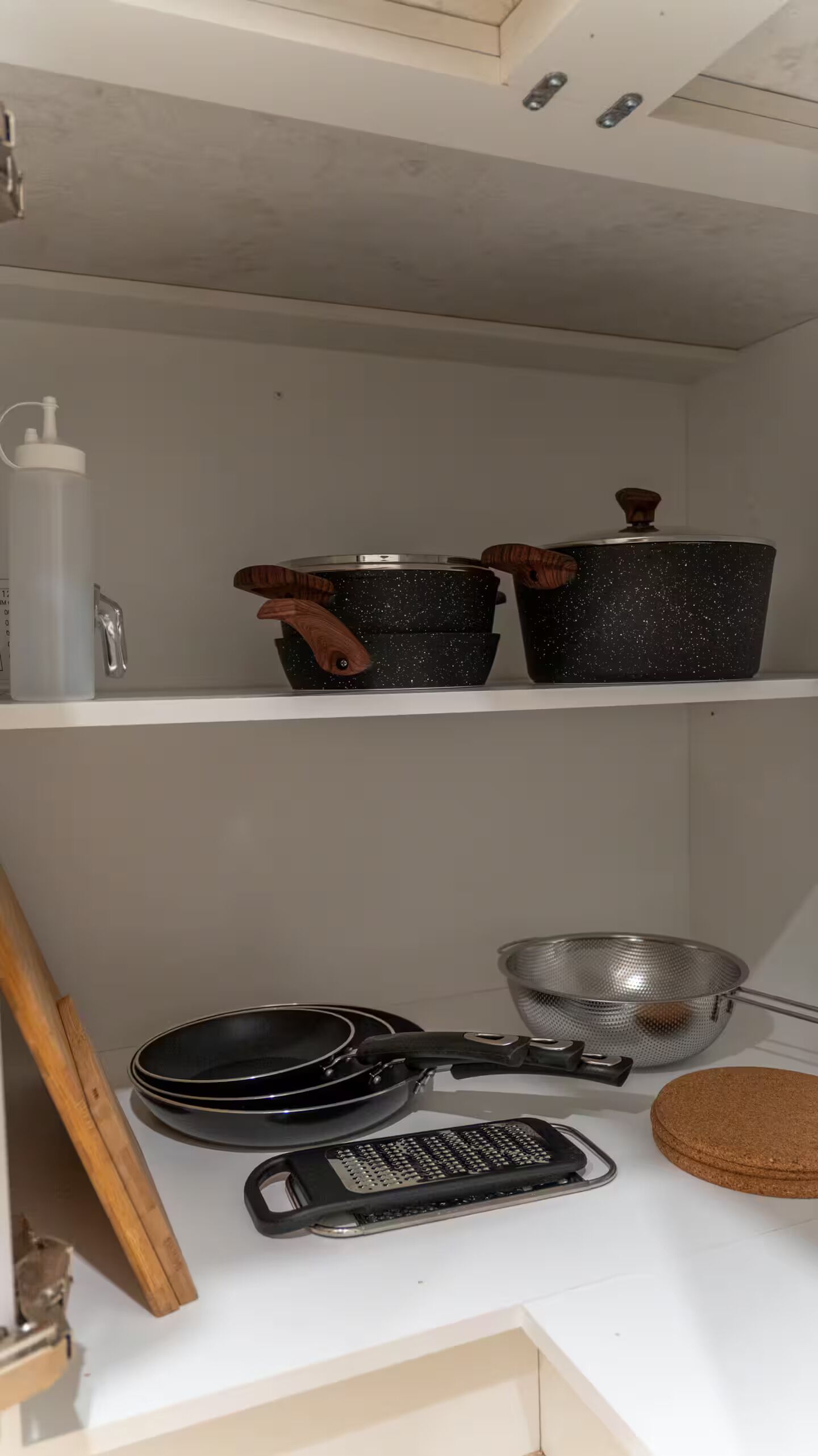 Een keuken met een wit aanrecht, een rasp, een koekenpan, een vergiet, en twee pannen met houten handgrepen.