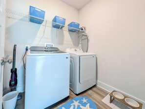 A laundry area with a washing machine, dryer, and a shelf with blue baskets. - NEW! 5BR WindMark | Golf Cart | Pet Friendly (Port St. Joe)