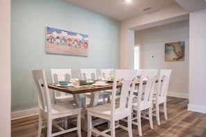 A dining area with a table set for six, white chairs, and a painting of beach huts on the wall. - NEW! 5BR WindMark | Golf Cart | Pet Friendly (Port St. Joe)