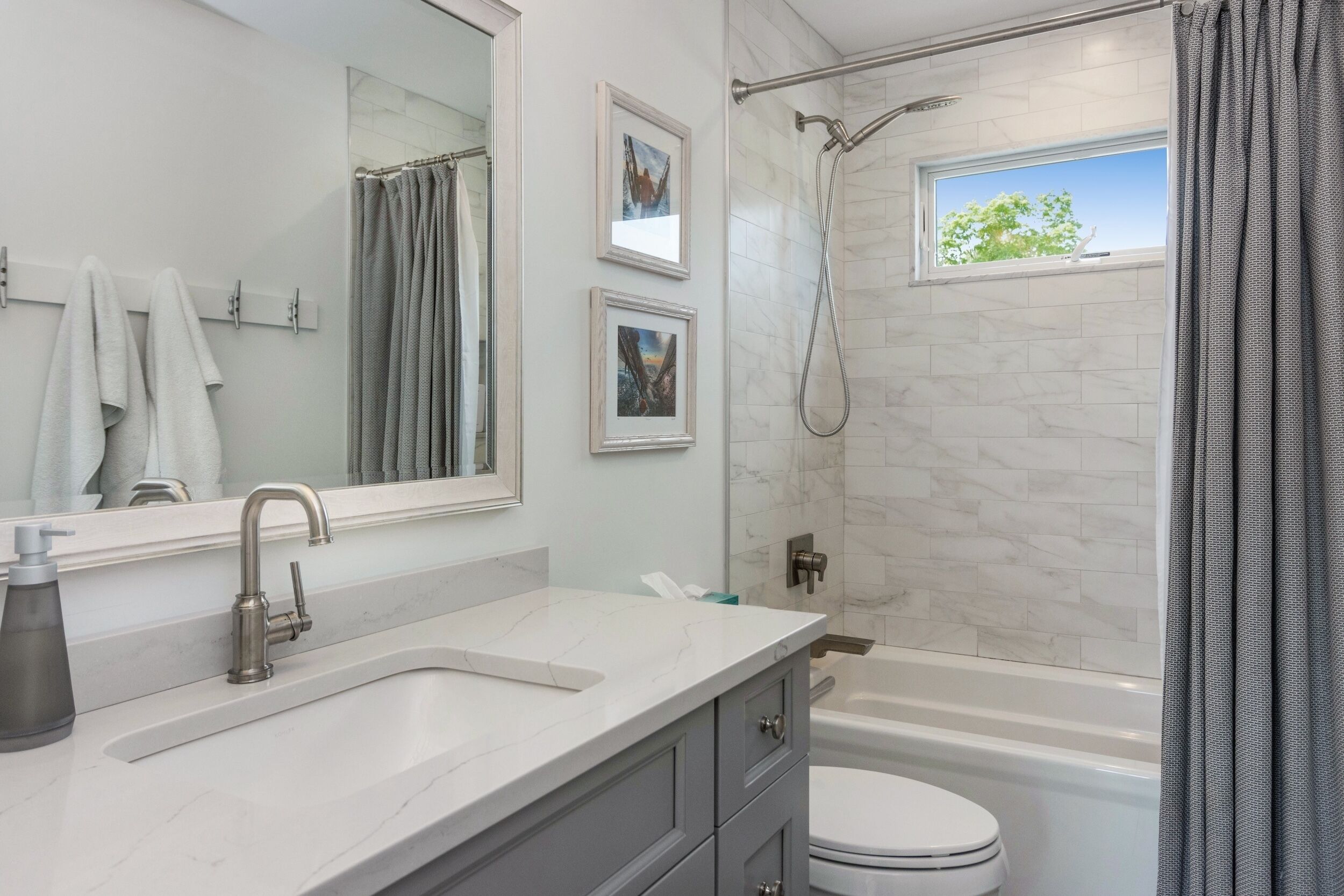 Une salle de bain équipée d’une baignoire, de toilettes, d’un lavabo et d’une douche. On y trouve des serviettes suspendues à un porte-serviettes, une fenêtre donnant sur des arbres et des tableaux encadrés accrochés au mur.