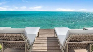 Eine Holzterrasse führt zu zwei Korbsesseln mit weißen Kissen, mit Blick auf ein klares, türkisblaues Meer.