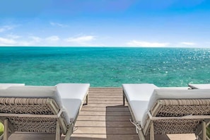 Eine Holzterrasse führt zu zwei Korbsesseln mit weißen Kissen, mit Blick auf ein klares, türkisblaues Meer.
