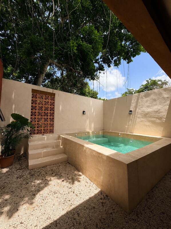 Un bain à remous rectangulaire installé à l’extérieur, avec des marches menant à l’intérieur, entouré d’un mur de couleur beige et d’un arbre.