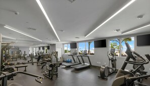 A modern gym with various exercise equipment, large windows, and a view of palm trees. - Sleek Miami 2BR | Free parking | Pool & Gym (Miami)