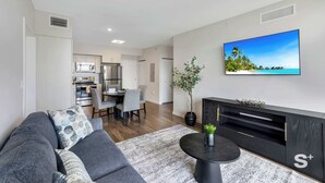 A modern living room with a sofa, coffee table, and TV. There is a dining area with chairs and a table in the background. - Sleek Miami 2BR | Free parking | Pool & Gym (Miami)