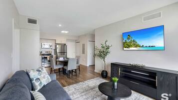Una sala moderna con sofá, mesa de centro y televisor. Se observa un comedor con sillas y una mesa al fondo.