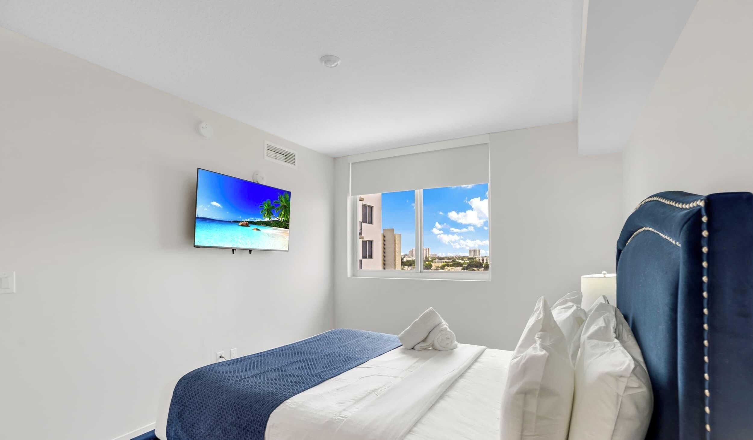 Habitación de hotel con cama, ventana con vista a la ciudad, televisor de pared mostrando una escena de playa y techo blanco.