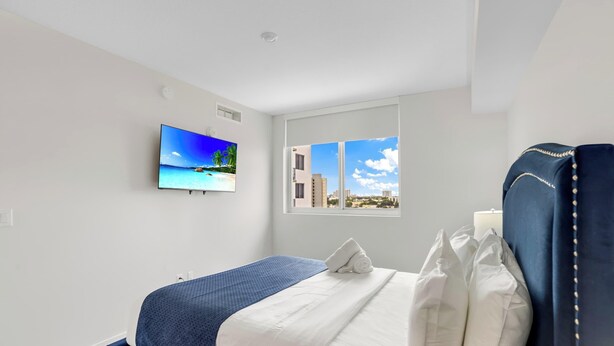 A hotel room with a bed, a window with a city view, a wall-mounted TV displaying a beach scene, and a white ceiling. - Sleek Miami 2BR | Free parking | Pool & Gym (Miami)