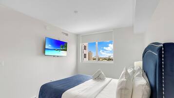Habitación de hotel con cama, ventana con vista a la ciudad, televisor de pared mostrando una escena de playa y techo blanco.